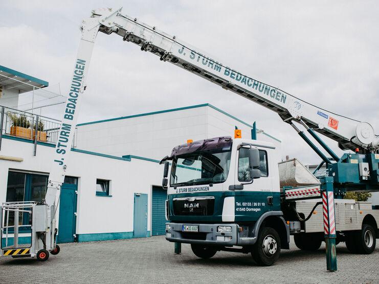 Mobiler Kran auf einem Betriebshof Ein mobiler Kranwagen von "J. Sturm Bedachungen" auf einem gepflasterten Hof vor einem modernen, weißen Gebäude. Der Ausleger des Krans ist weit ausgefahren.