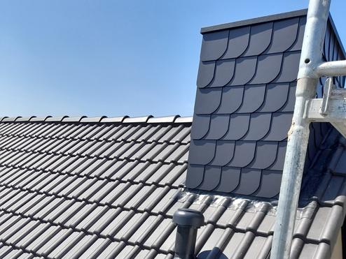 Detailansicht eines modernen Daches mit Belüftungselementen Ein Teil eines grauen Ziegeldaches mit einer modernen Dachentlüftung und einer Schornsteinverkleidung. Eine Metallleiter ist an der Seite sichtbar, unter klarem blauem Himmel.