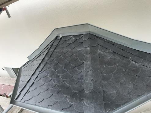 Schiefereindeckung auf einer Dachkante Detailansicht der schieferten Dachschindeln auf einer Dachkante, eingerahmt von grauen Metallleisten an den Seiten. Die Schindeln sind in einem dekorativen Fischschuppenmuster angeordnet.