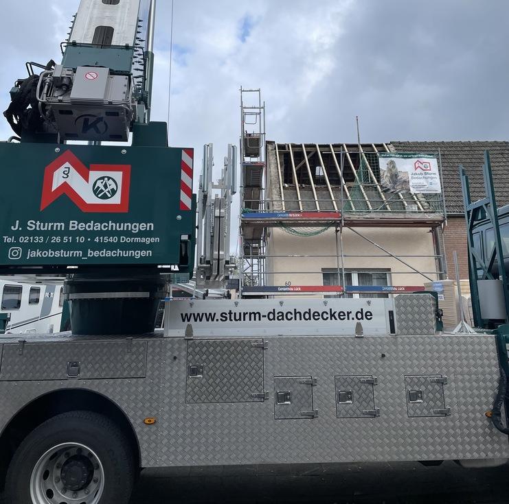 Baustelle mit Kran und Gerüst an einem Haus Ein großer, mobil montierter Kran an einer Baustelle, der vor einem Haus mit Gerüst steht. Der Himmel ist bewölkt.