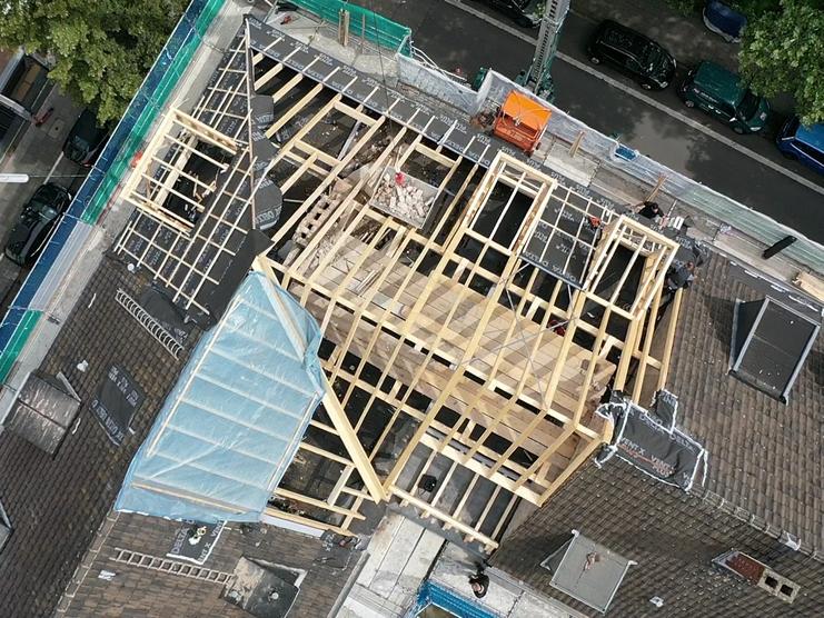 Luftaufnahme einer Baustelle mit Holzkonstruktion Drohnenansicht einer aktiven Baustelle, die den Aufbau eines großen Holzrahmens auf einem Dach zeigt. Bauarbeiter sind sichtbar. Umgeben von städtischer Umgebung und parkenden Autos.