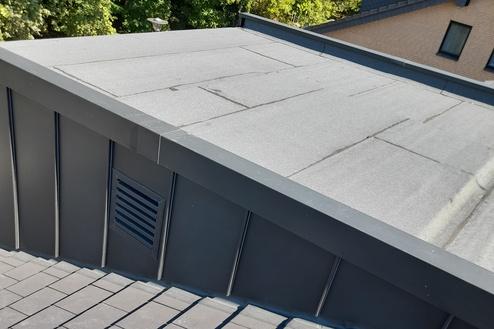 Modernes Flachdach mit dunkler Verkleidung Eckansicht eines modernen Flachdachs mit dunkelgrauer Verkleidung und Dachbahnen, umrahmt von einem grünen Laubwald im Hintergrund.