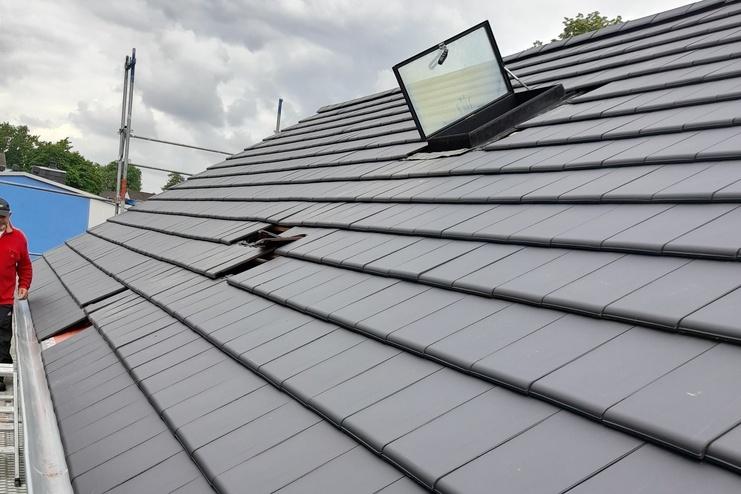 Neues dunkelgraues Dach mit Dachfenster Frisch installierte dunkelgraue Dachziegel auf einem geneigten Dach mit einem offenen Dachfenster und Holzwerkzeug darauf. Bedeckter Himmel im Hintergrund, Bauarbeiter teilweise sichtbar.