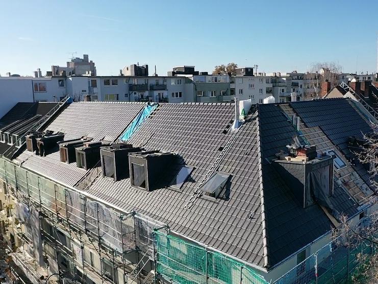 Städtische Dachlandschaft mit Renovierungsarbeiten Luftaufnahme von mehreren zusammenhängenden Dächern mit grauen Ziegeln, umgeben von modernen Gebäuden. Einige Dächer haben offene Baustellen mit Schutznetzen und Gerüsten.