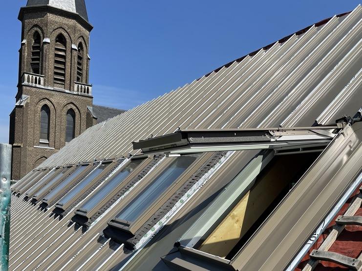 Installation von Dachfenstern auf einem Schieferdach Ansicht von oben auf ein Schieferdach mit mehreren frisch installierten Dachfenstern. Im Hintergrund ragt der Turm einer Kirche empor. Klare Sicht bei sonnigem Wetter.