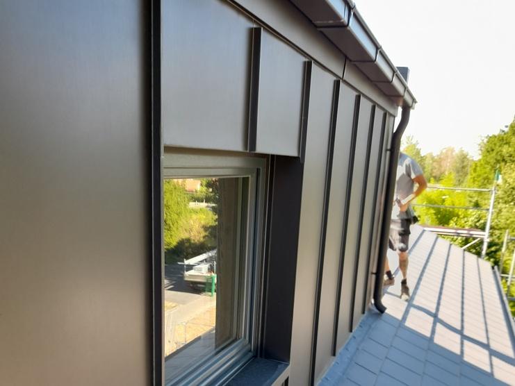 Arbeiter auf einem modernen Flachdach Ein Arbeiter auf einem Flachdach mit dunkler Metallverkleidung und -fenstern. Er bewegt sich entlang des Dachs, das von einem sonnigen Tag beleuchtet wird, mit Blick auf grüne Umgebung.