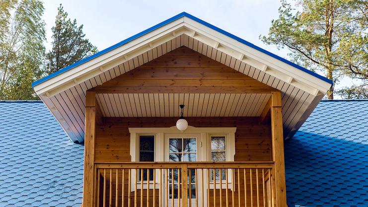 Blick auf einen Holzbalkon mit zwei Fenstern unter einem giebelförmigen Dach auf einer blauen Schindelfassade.